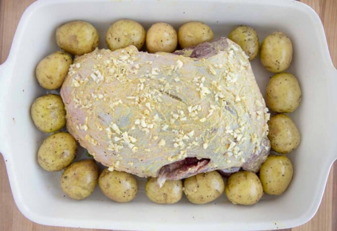 leg of lamb coated with mustard and chopped garlic in a pan with baby potatoes around the roast