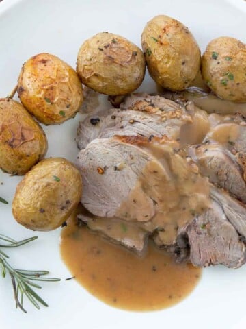 sliced leg of lamb with gravy along side whole baby potatoes and a sprig of rosemary on a white plate