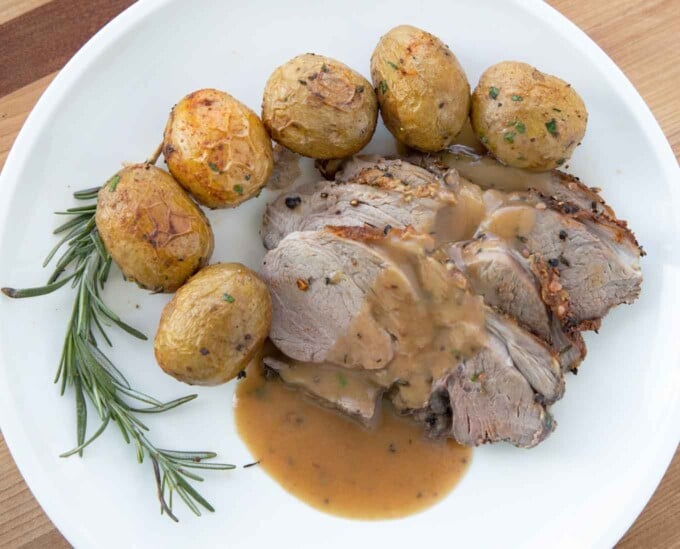 sliced leg of lamb with gravy along side whole baby potatoes and a sprig of rosemary on a white plate