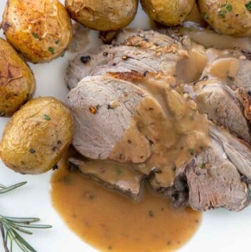 sliced leg of lamb with gravy along side whole baby potatoes and a sprig of rosemary on a white plate