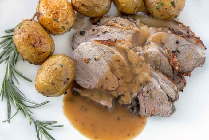 sliced leg of lamb with gravy along side whole baby potatoes and a sprig of rosemary on a white plate