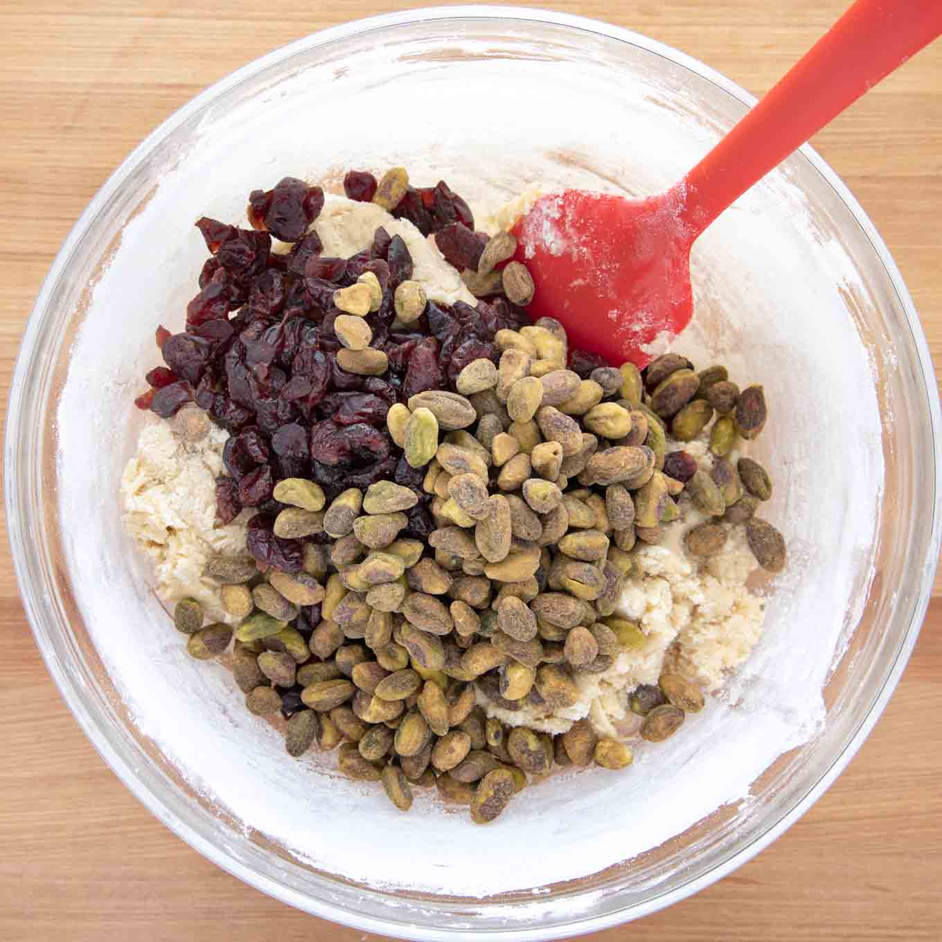 pistachios and craisins added to  dough in a glass bowl with a red spatula