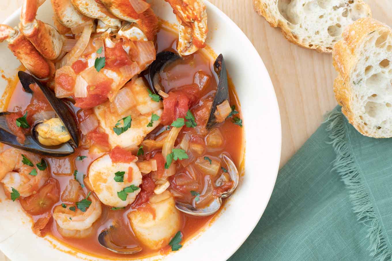 overhead shot of cioppino in a white bowl next to slices of italian bread and a green napkin