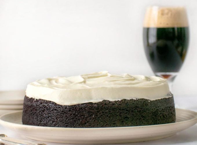 Whole frosted Guinness chocolate cake on a cream colored platter with a pint of Guinness behind it