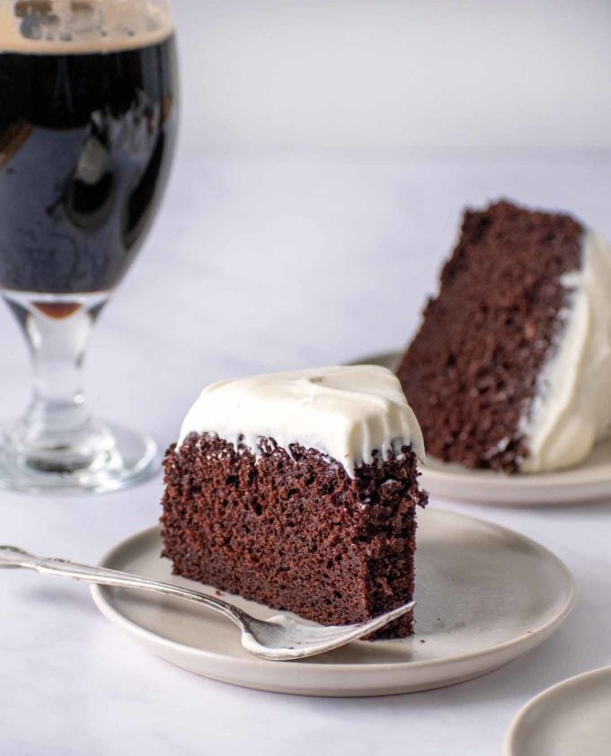 slice of Guinness chocolate cake with a fork on a cream colored plate with a pint of Guinness and another slice of cake behind it