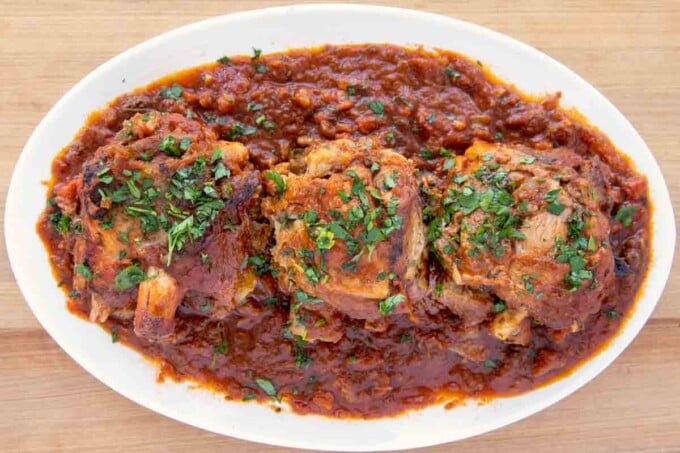 three pork shanks on a bed of tomato sauce on a white oval platter