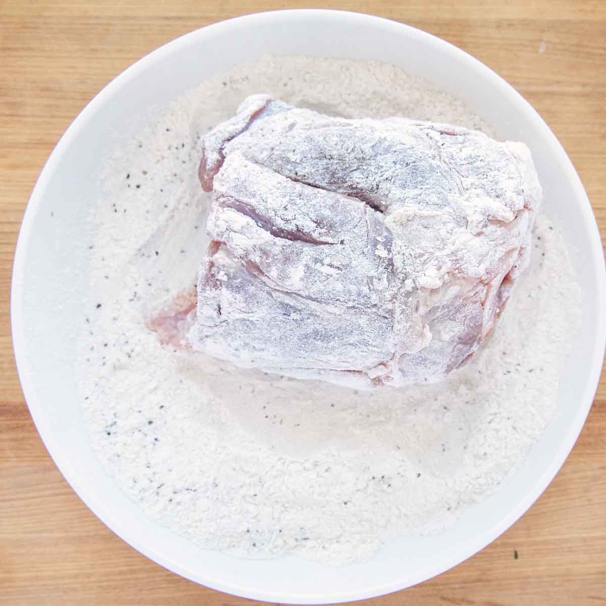 floured pork shank in a white bowl of seasoned flour