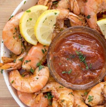 partial overhead view of peel and eat shrimp with cocktail sauce and lemons in a white bowl