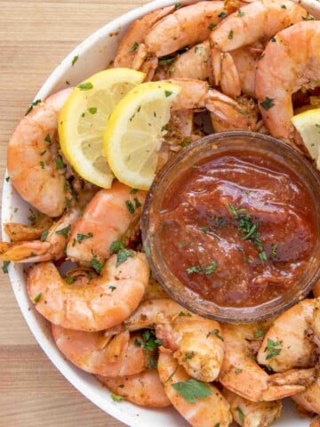 partial overhead view of peel and eat shrimp with cocktail sauce and lemons in a white bowl