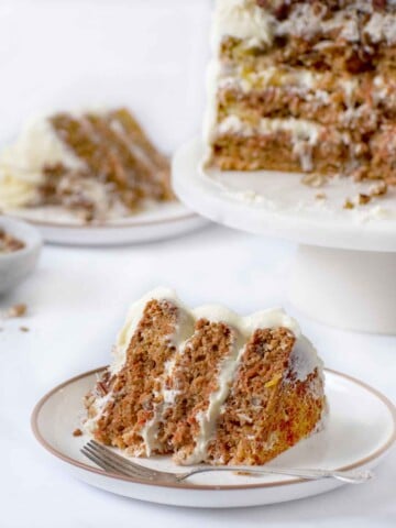 slice of carrot cake on a plate with a fork with an other slice and the remainder of the whole cake on a pedestal in the background