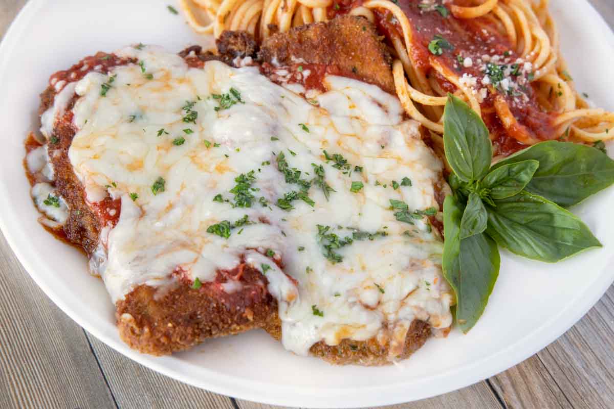 close up of eggplant parm with a side of linguine on a white plate with a sprig of basil