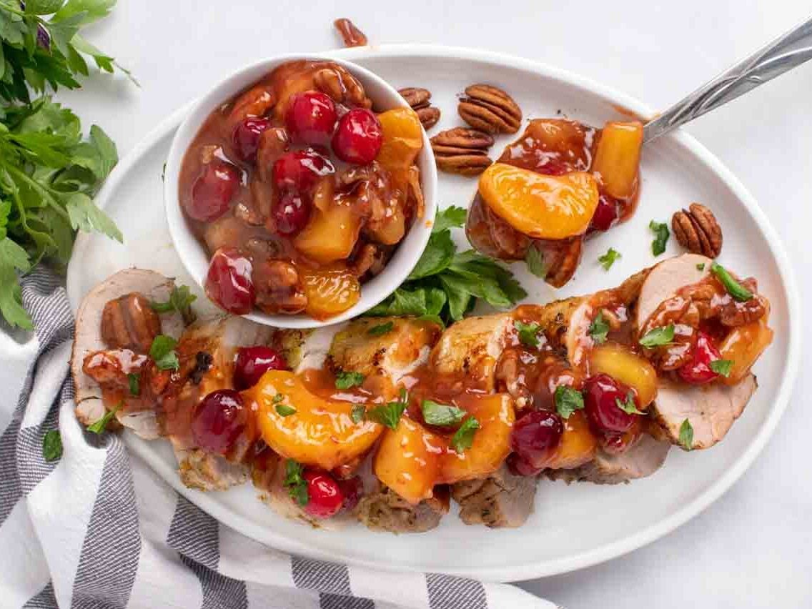 overhead view of slices of peppered pork loin topped with the sweet chili pecan fruit sauce with a bowl fo the sauce on a white plate