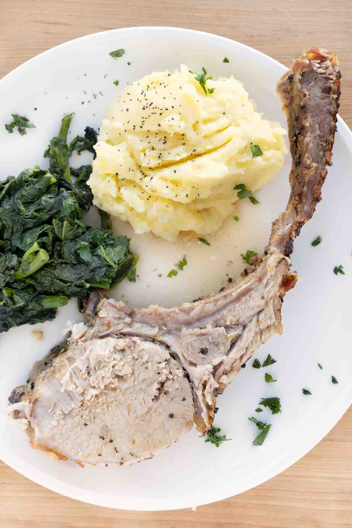 tomahawk pork chop on a white plate with mashed potatoes and greens