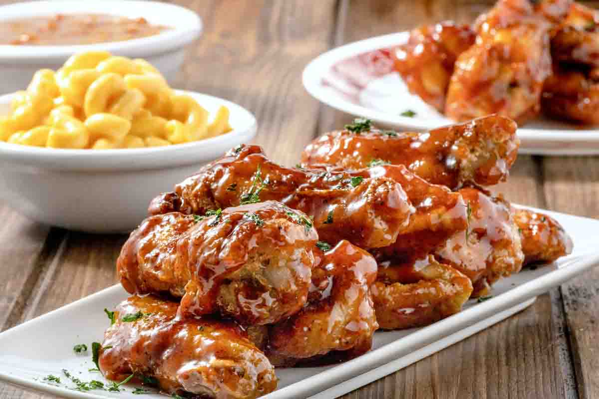 honey bbq wings on a white platter with more wings and mac and cheese in the background