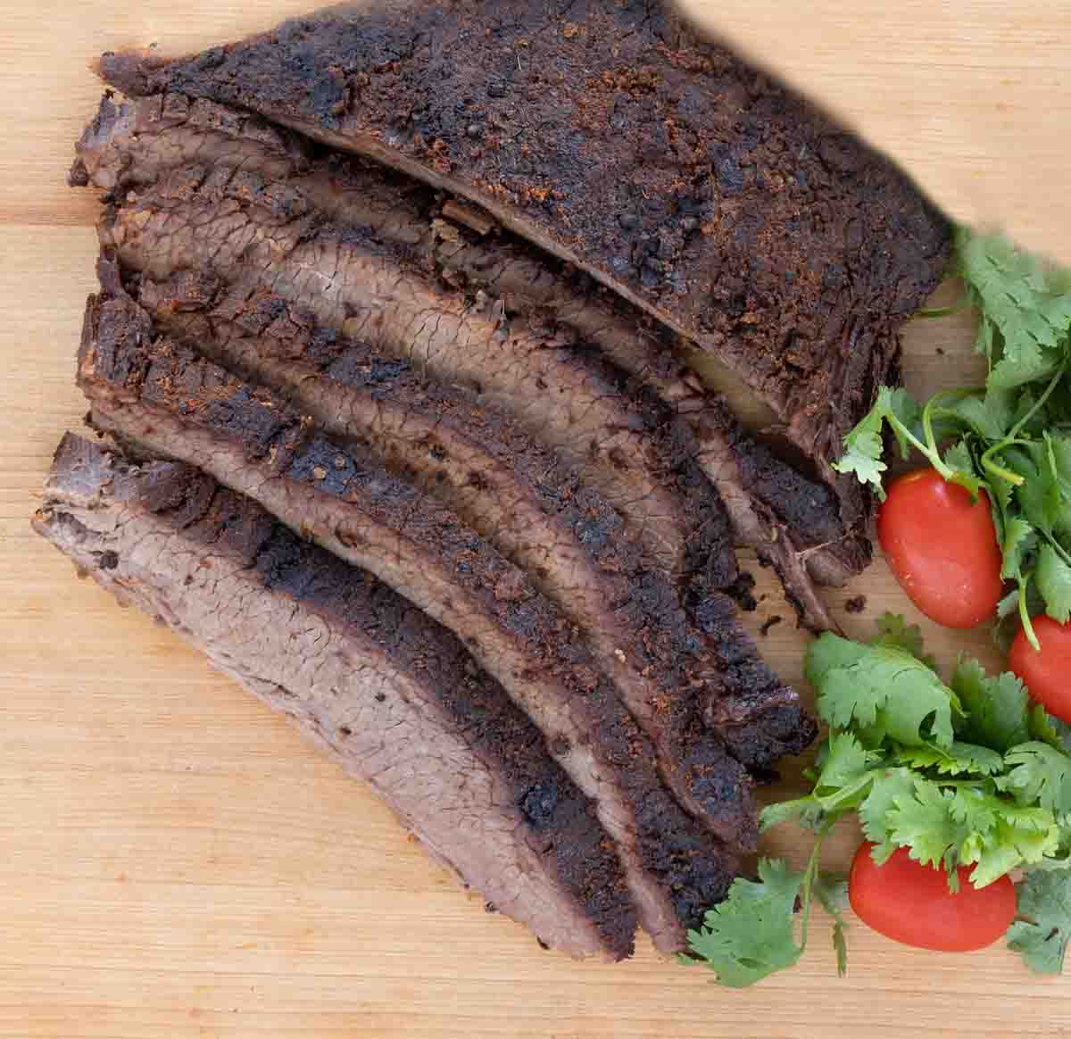 sliced dry rub roasted brisket with cilantro and tomatoes on wooden cutting board