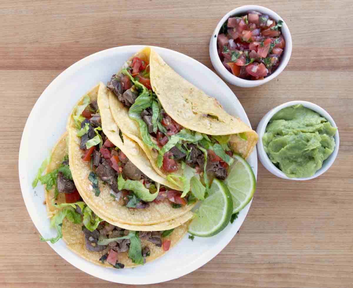 3 brisket tacos on a white platter with dishes of guacamole and salsa next to it