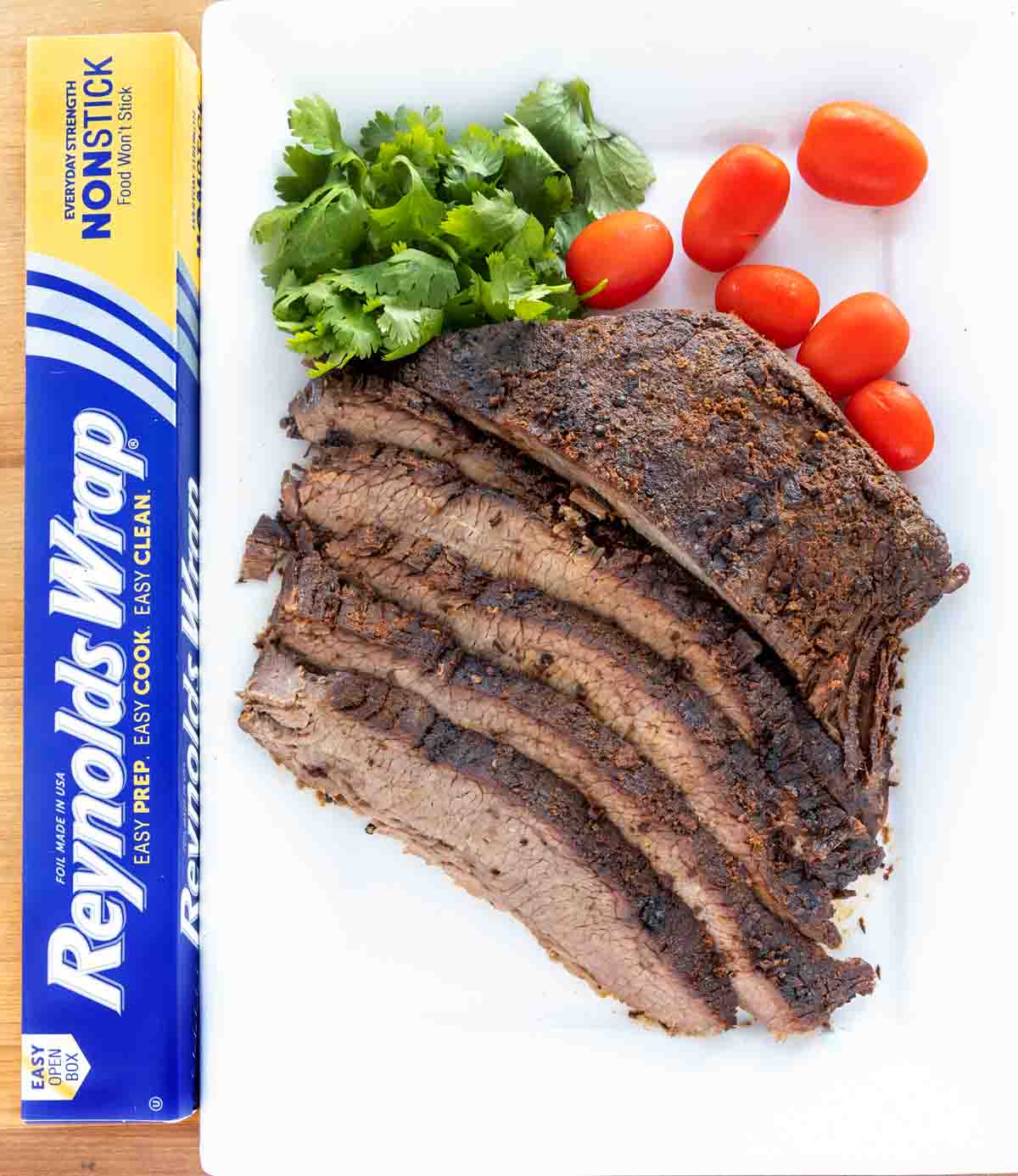 sliced dry rub roasted brisket with cilantro and tomatoes on on a white platter next to a box of reynolds foil