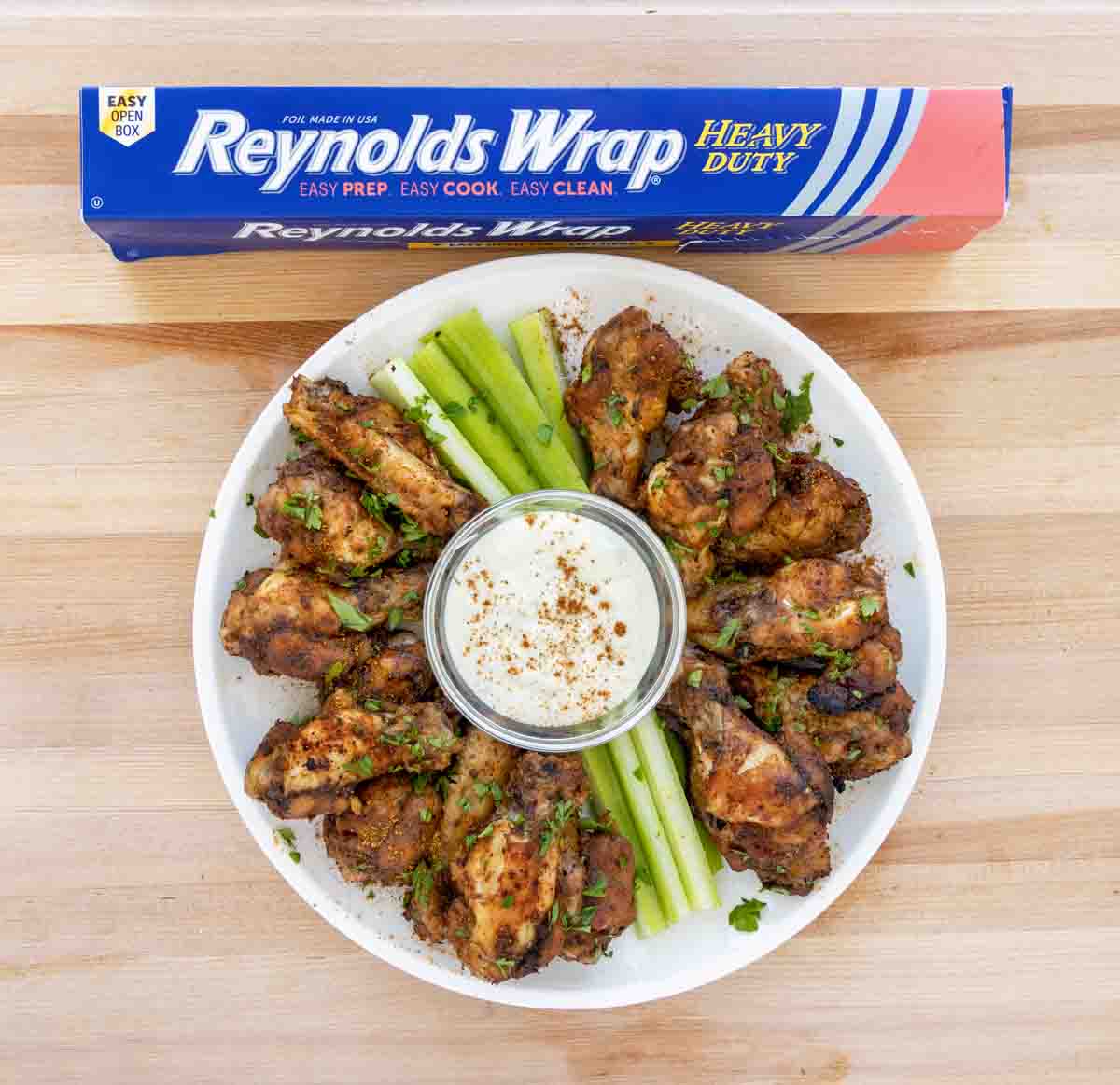 Cajun chicken wings on a white plate with celery and bleu cheese dressing next to a box of Reynolds foil
