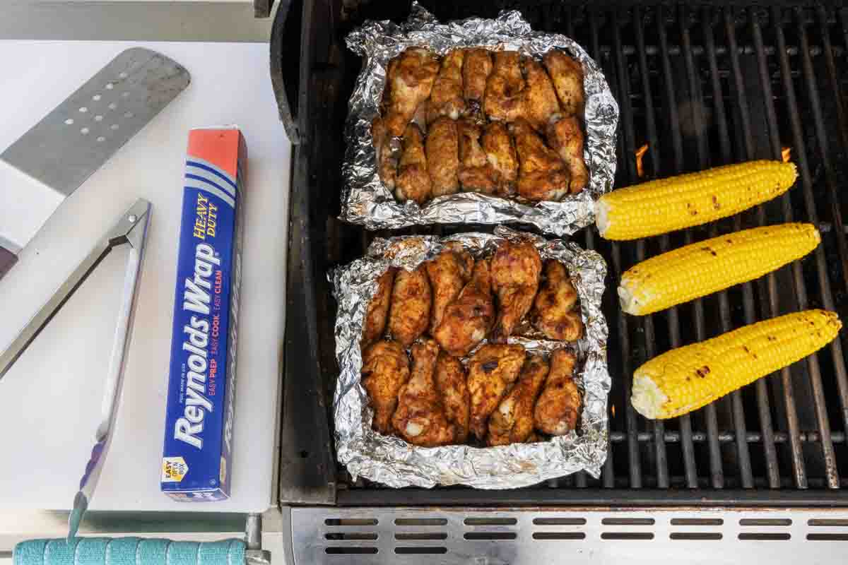 chicken wings in foil pouches on grill next to corn on the cob 