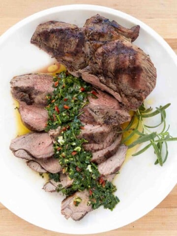 sliced and unsliced grilled leg of lamb on a white plate with chimichurri sauce.