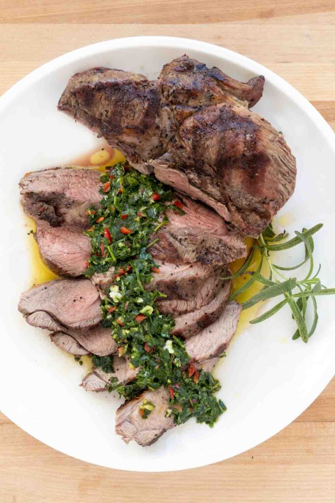 sliced and unsliced grilled leg of lamb on a white plate with chimichurri sauce.