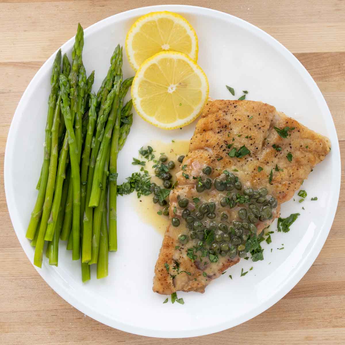 pan seared rockfish with a lemon caper sauce on a white plate with asparagus and lemon circles