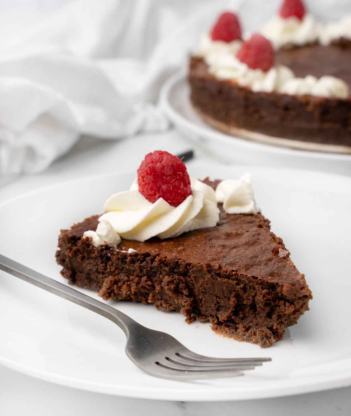 slice of flourless chocolate cake on a white plate topped with whipped cream and raspberries