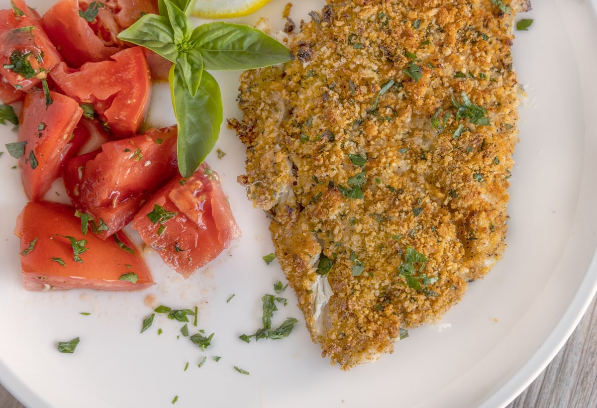 baked panko parmesan flounder on a white plate with lemons and tomatoes