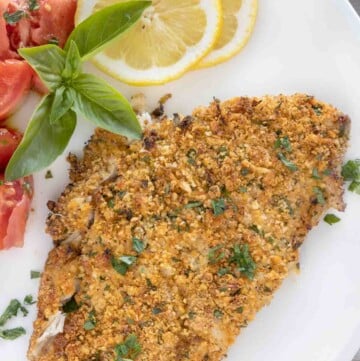 baked panko parmesan flounder on a white plate with lemons and tomatoes