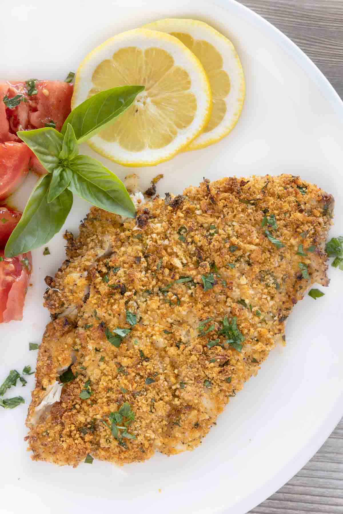 garlic parmesan flounder on a white plate with lemons and tomatoes