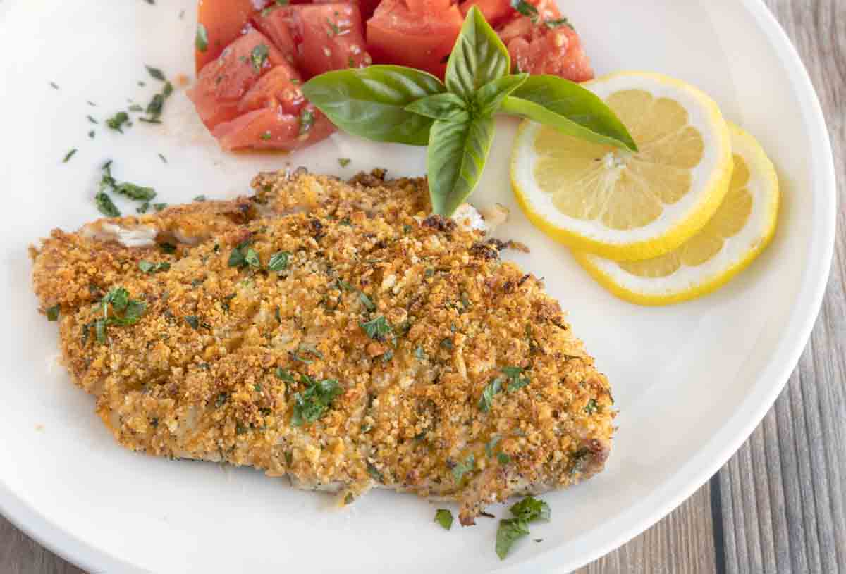 baked panko parmesan flounder on a white plate with lemons and tomatoes