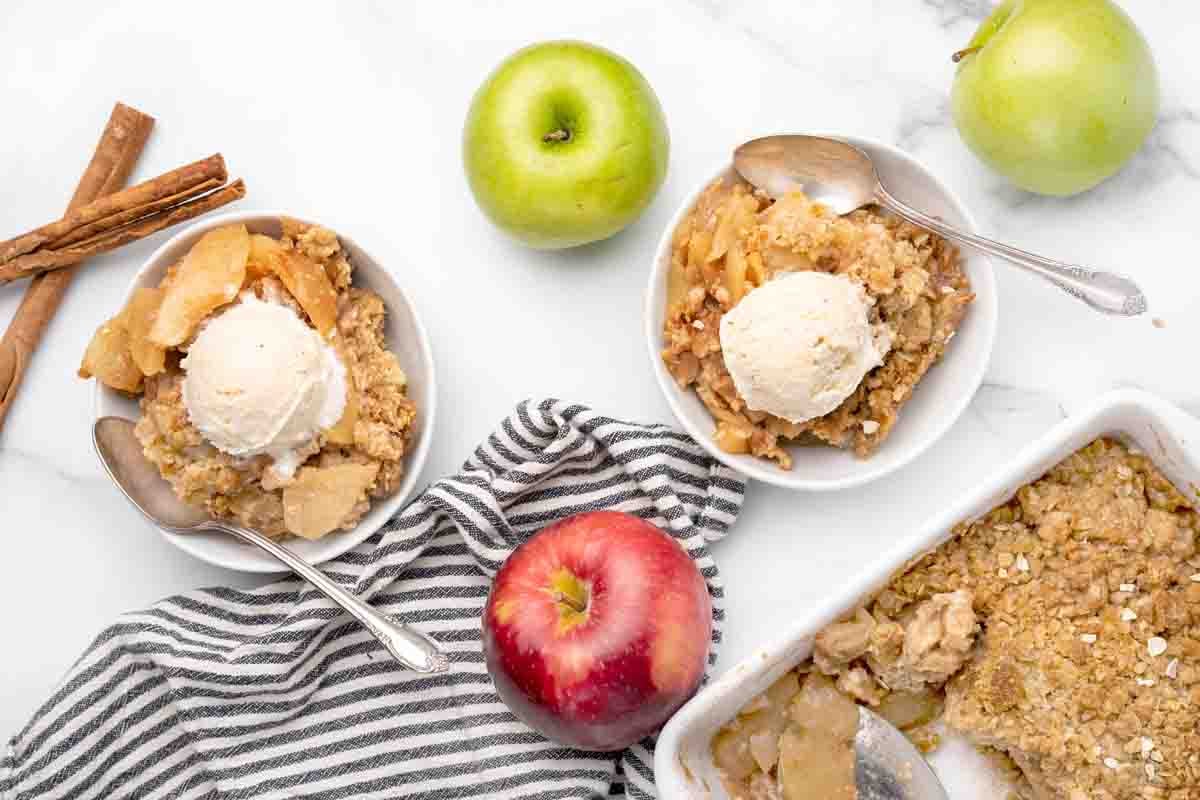 white bowls of apple crisp with ice cream next to whole red and green apples