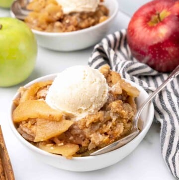 white bowl of apple crisp with ice cream next to whole red and green apples