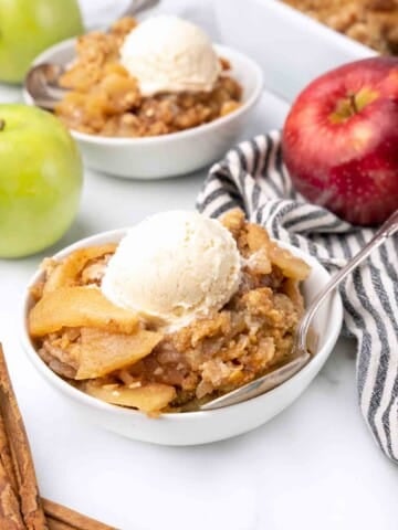 white bowl of apple crisp with ice cream next to whole red and green apples