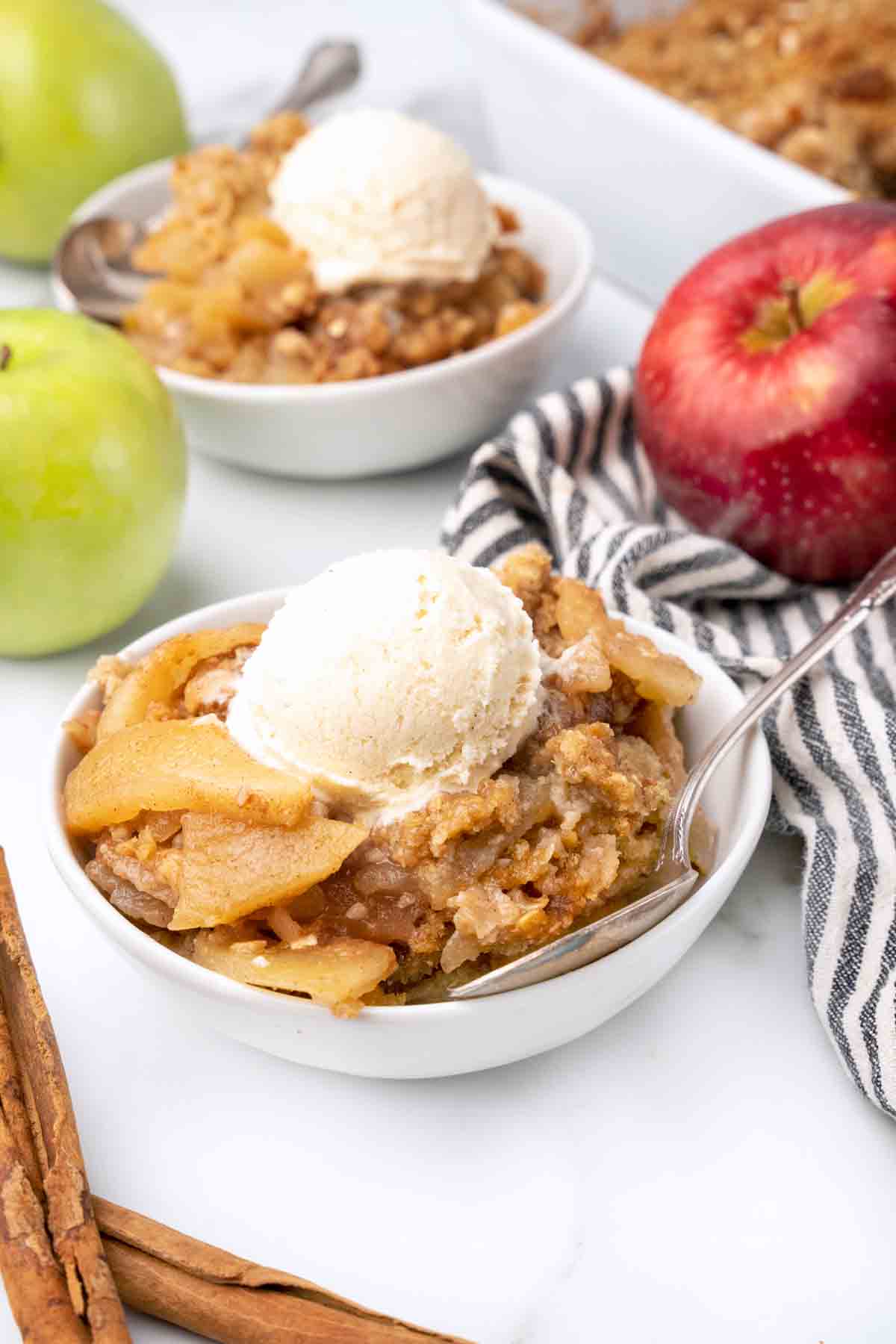 white bowl of apple crisp with ice cream next to whole red and green apples