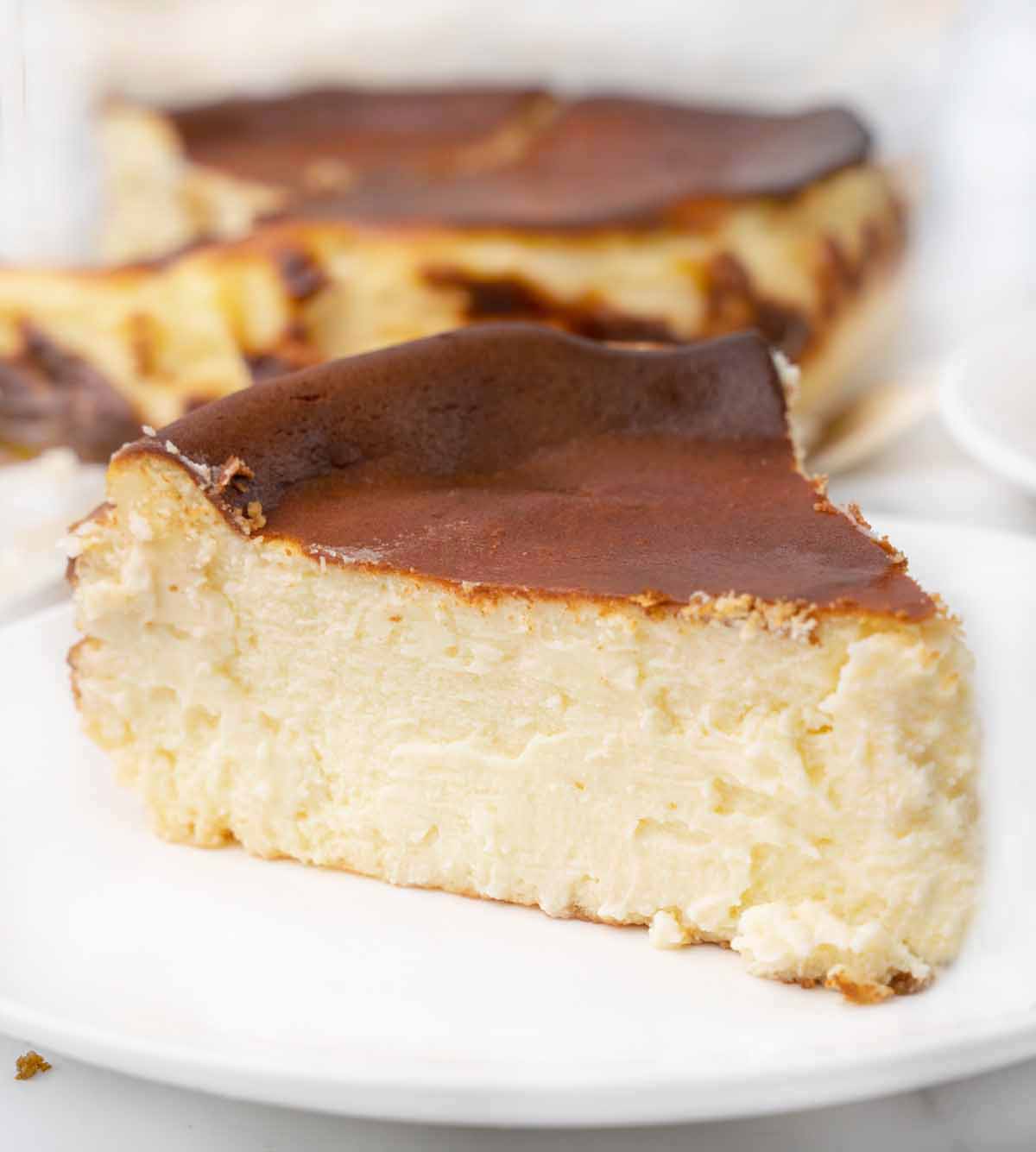 slice of basque cheesecake on a white plate with the rest of the cake behind it