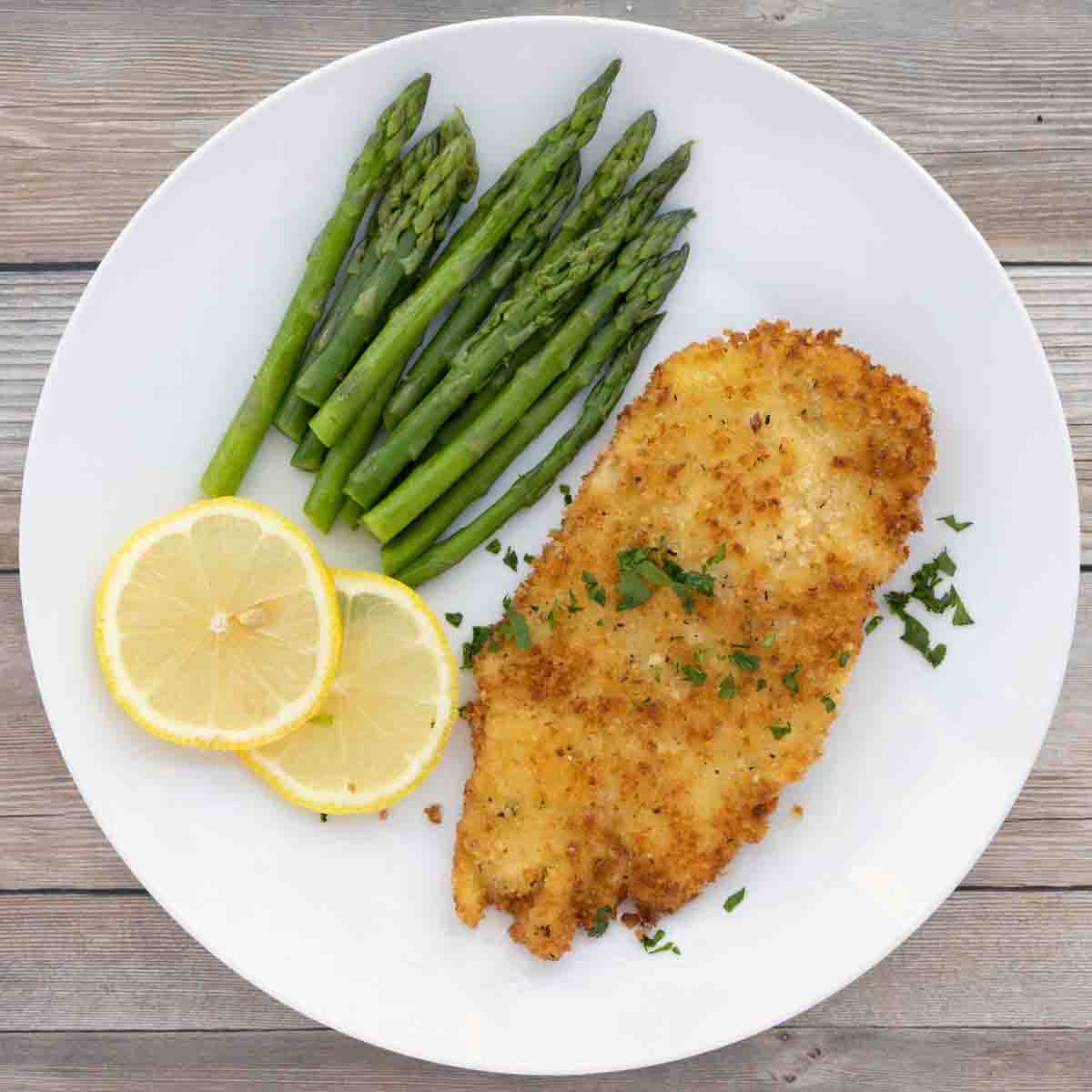 Chicken schnitzel on a white plate with lemon circles and asparagus.