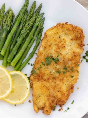 Chicken schnitzel on a white plate with lemon circles and asparagus.