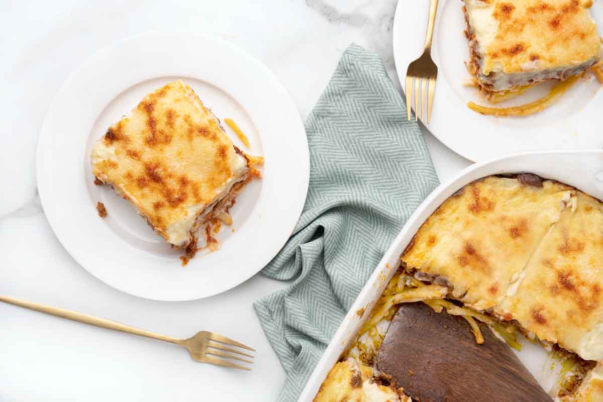 overhead view of slices of pastitsio on white plates next to a pan of pastitsio