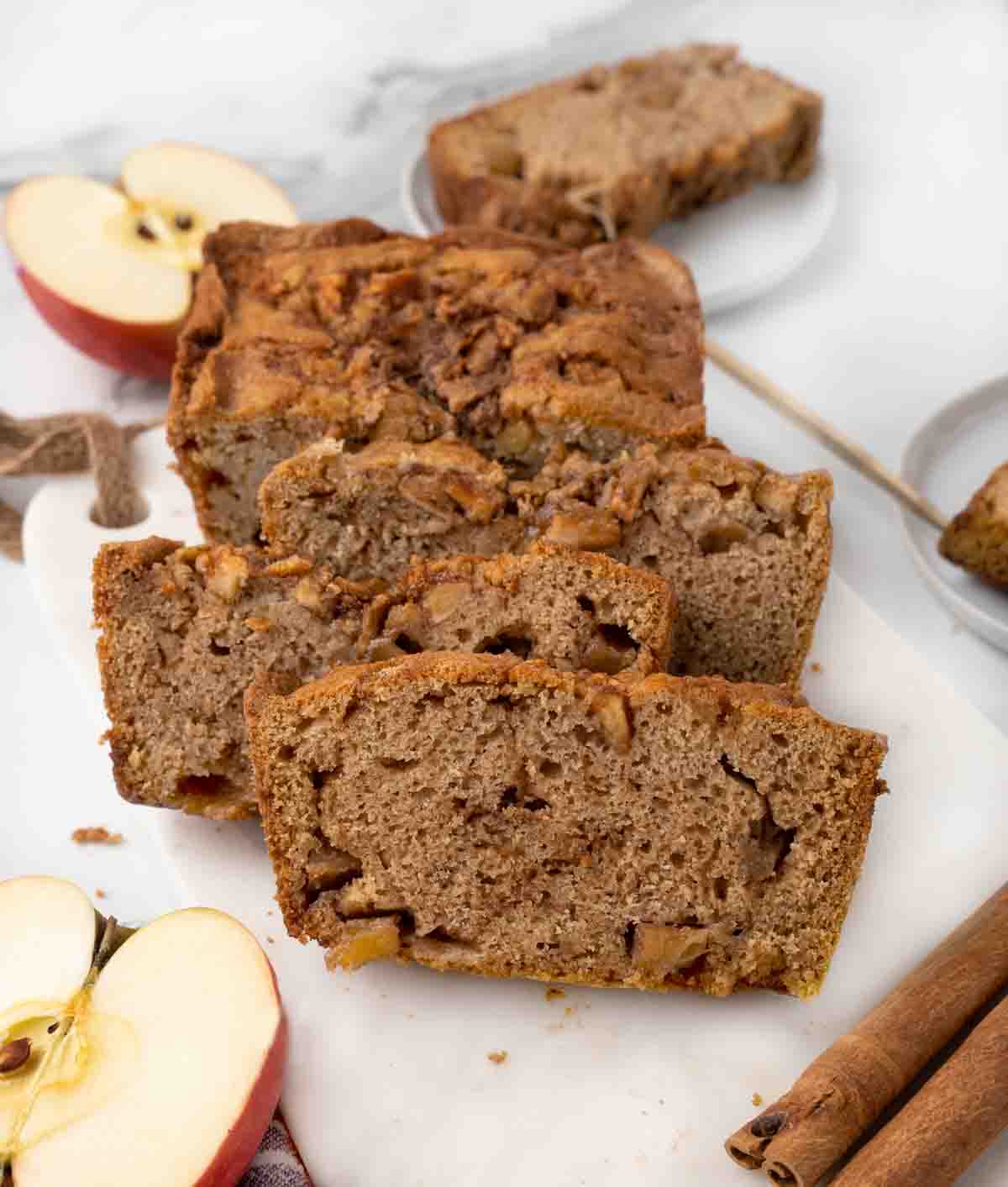 apple bread sliced with apples and cinnamon sticks on a white cutting board.
