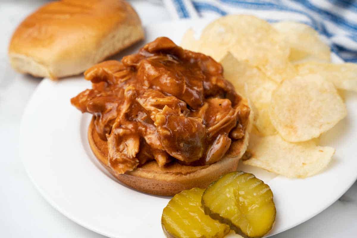 BBQ chicken on bun with potatoes chips and pickles on the side.