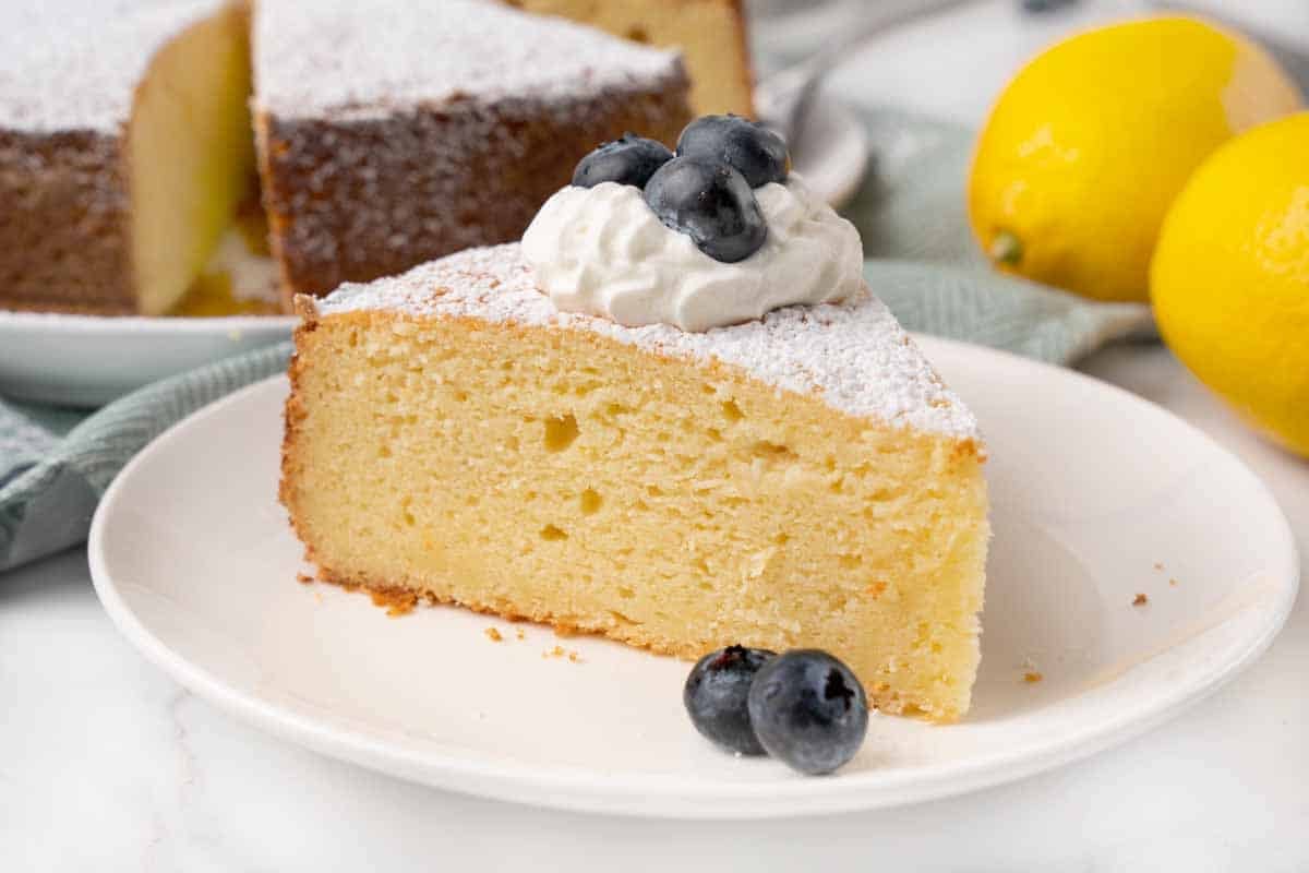 Slice of lemon ricotta cake on a white plate with whipped cream and blueberries.