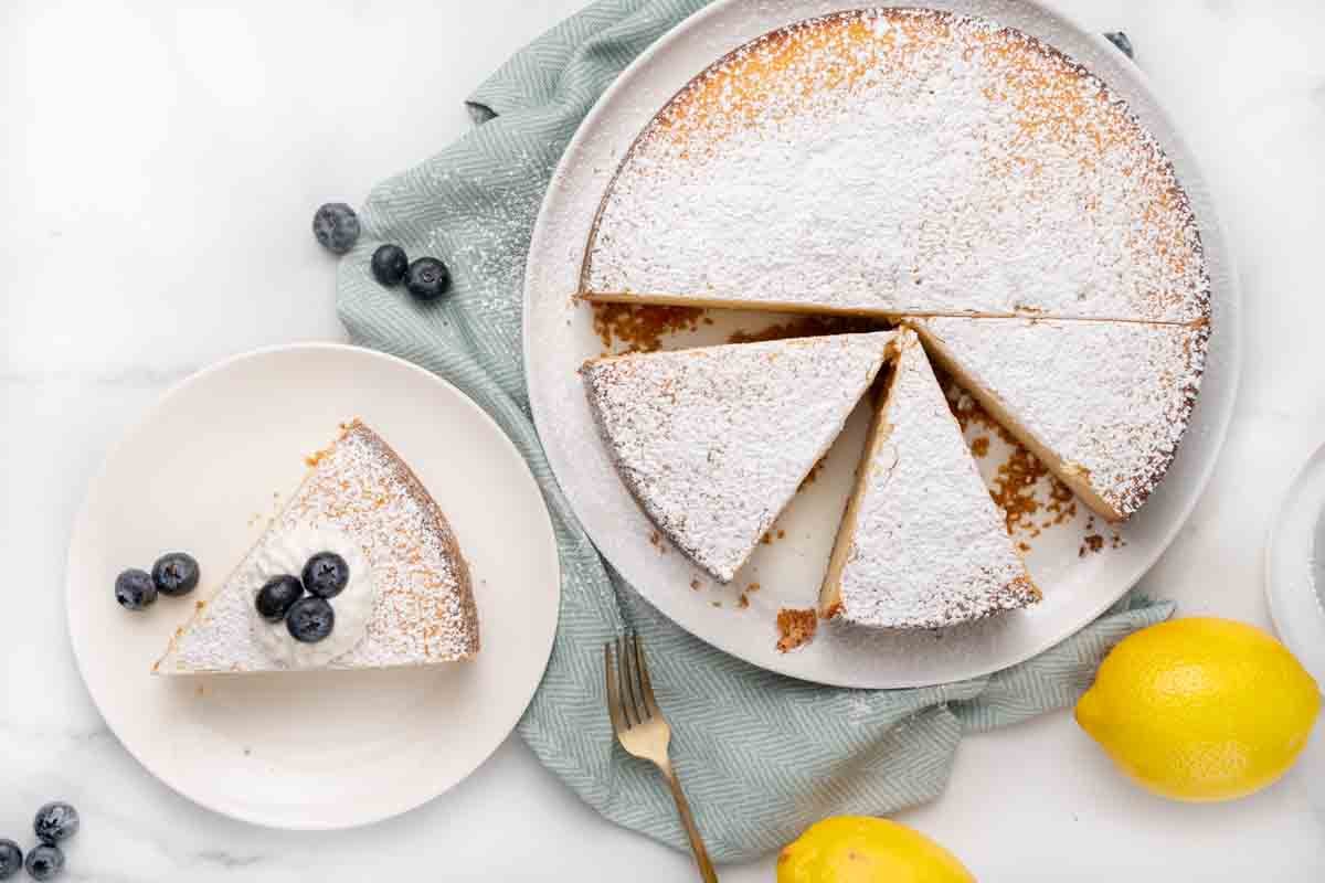 Sliced lemon ricotta cake on cake platter with one slice on a white plate.
