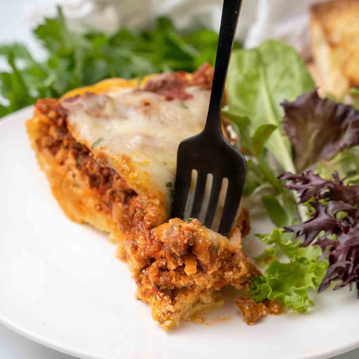 black fork in a slice of spaghetti pie on a white plate with salad.