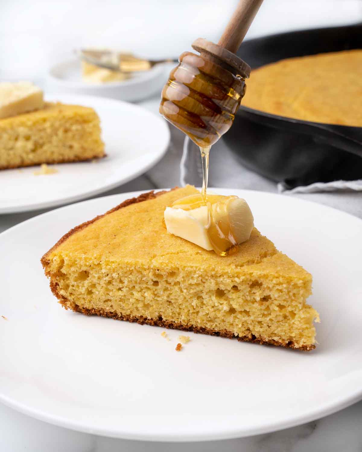 slice of skillet cornbread with pat of butter and honey on a white plate.