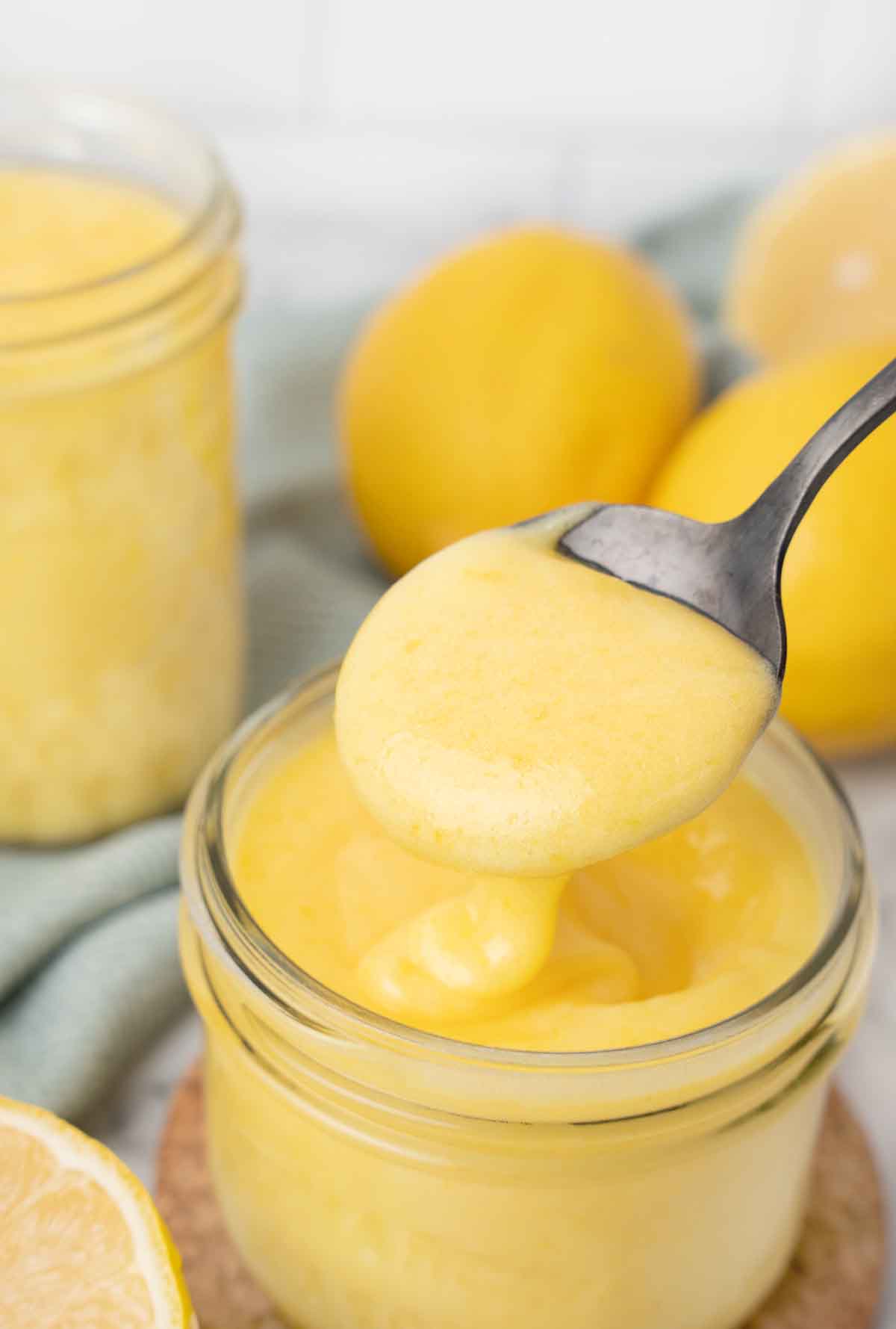 spoon in a jar of lemon curd, with lemons and another jar in the background.