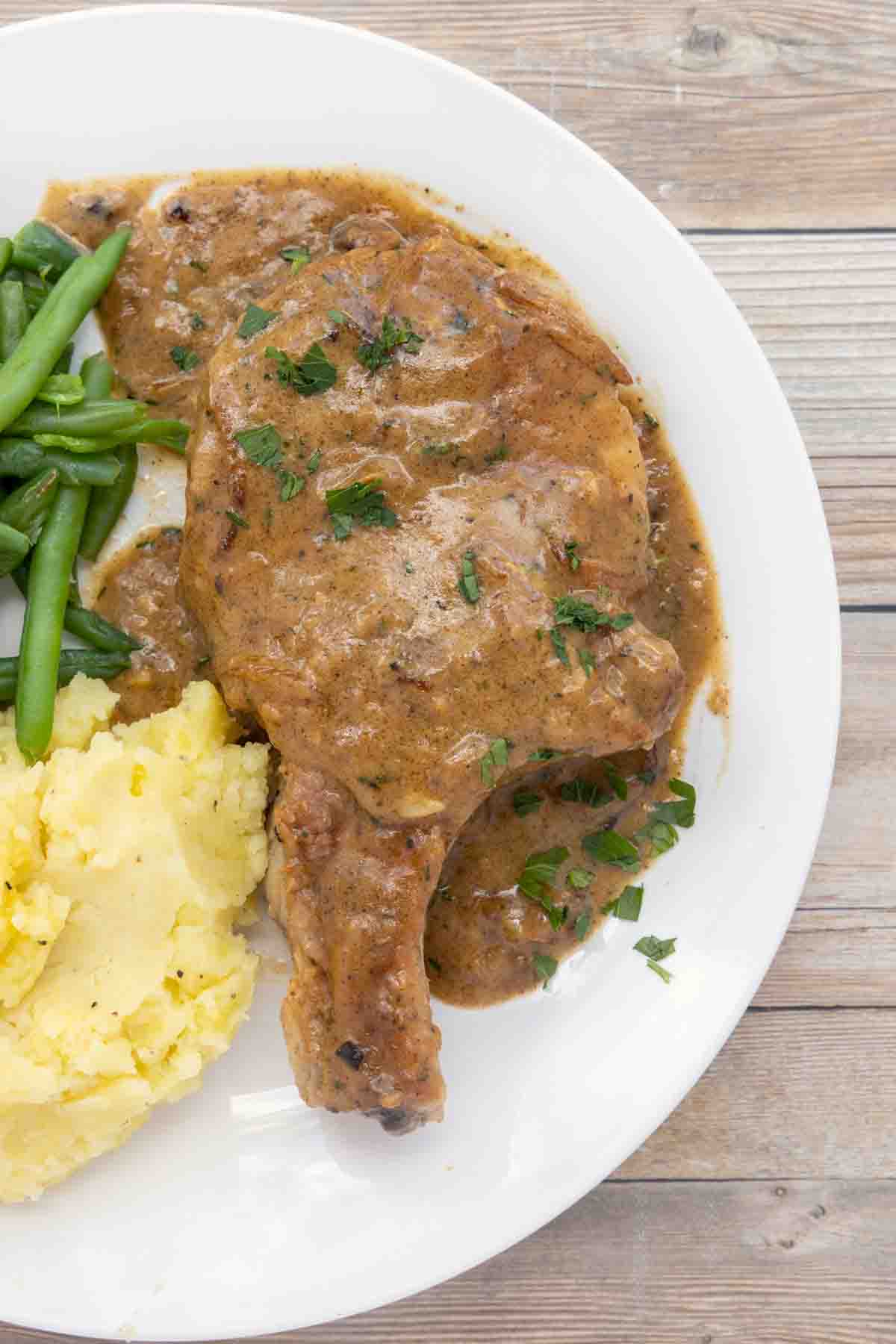 smothered pork chop on white plate with mashed potatoes and green beans.