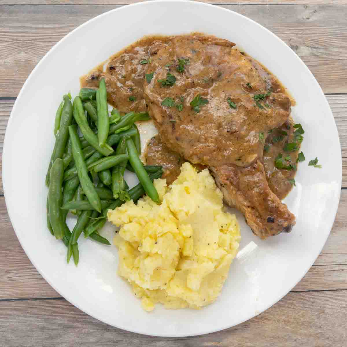 smothered pork chop on white plate with mashed potatoes and green beans.