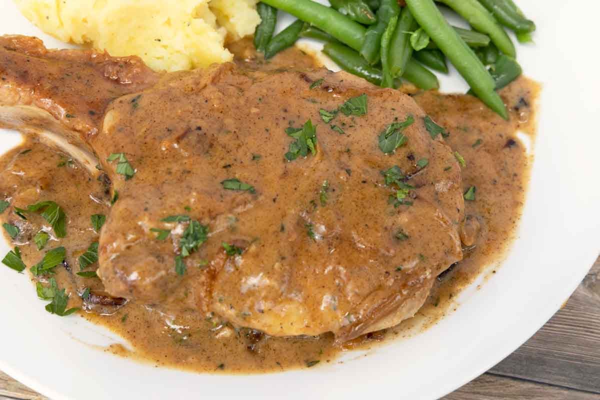 smothered pork chop on white plate with mashed potatoes and green beans.