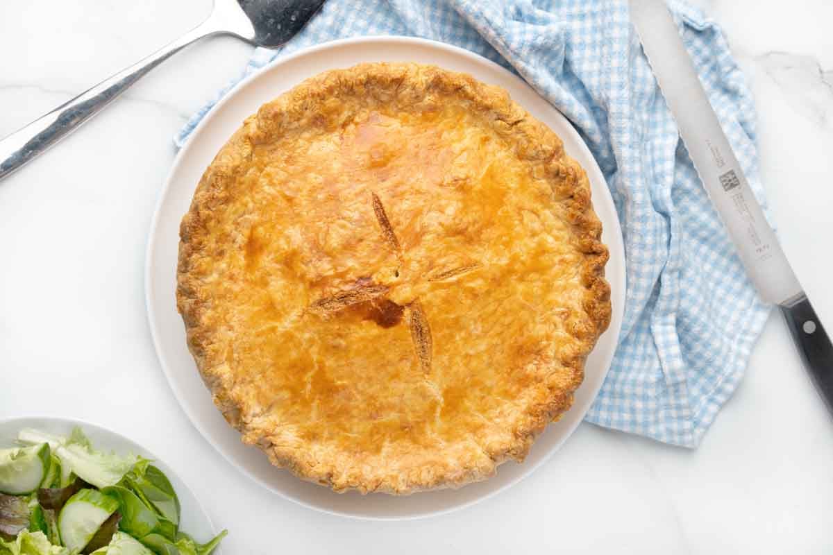 Whole Pizza Rustica with knife, blue checkered dishcloth and pie server on the side.